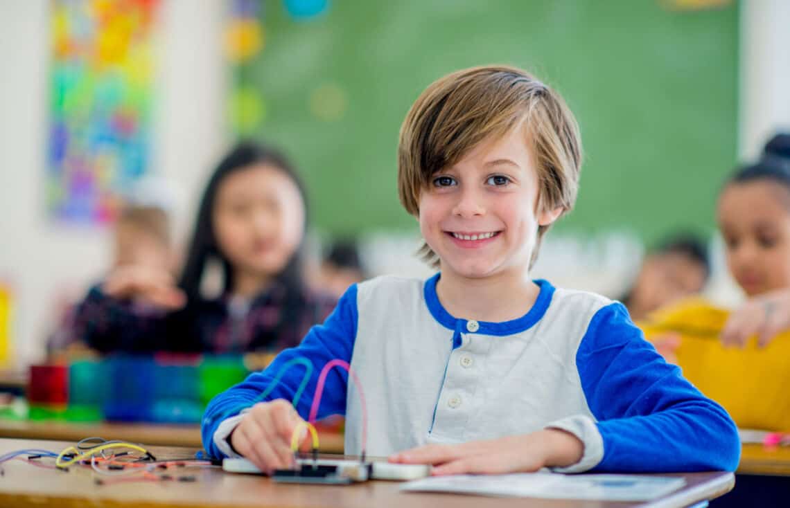 lächelnder weißer Junge sitzend in der Schule mit Kabeln im Physikunterricht