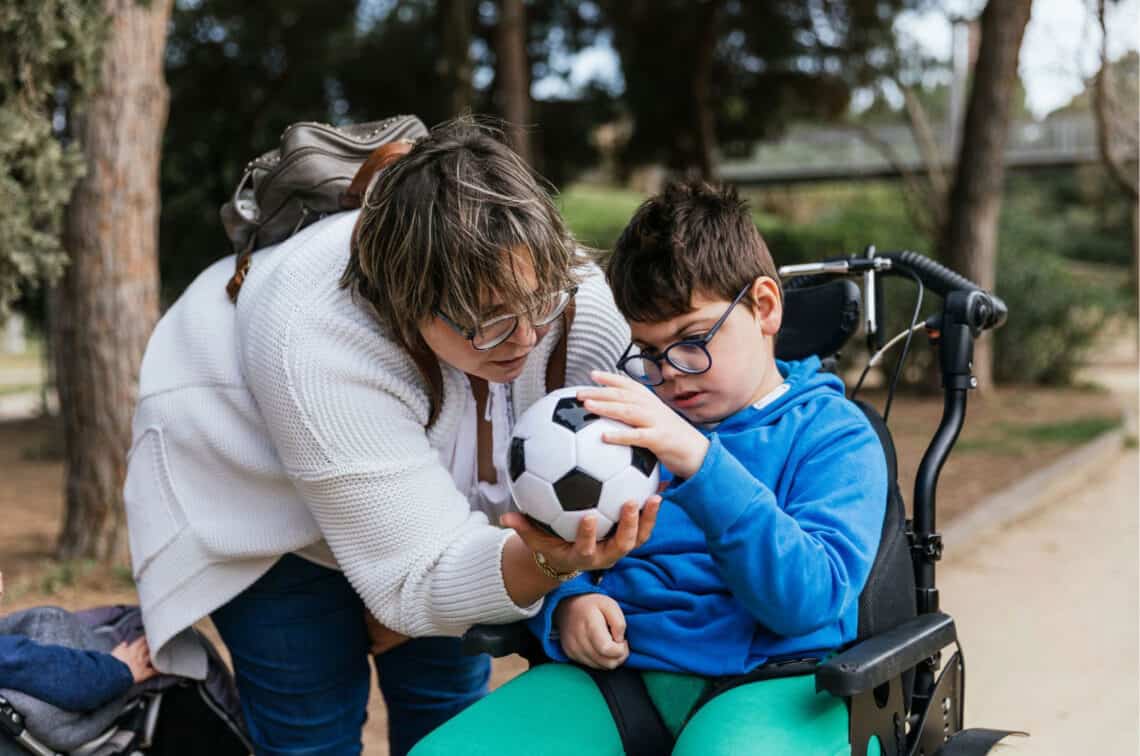 ein Junge im Rollstuhl in Park, dem eine Frau einen Fußball gibt