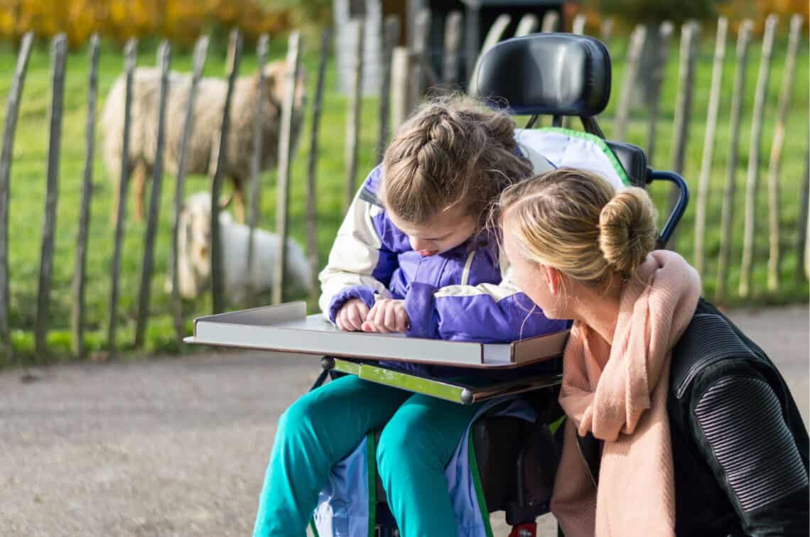 Mädchen im Rollstuhl, das zeichnet, daneben kniet eine Frau und spricht mit ihr, Wiese mit Schafen im Hintergrund