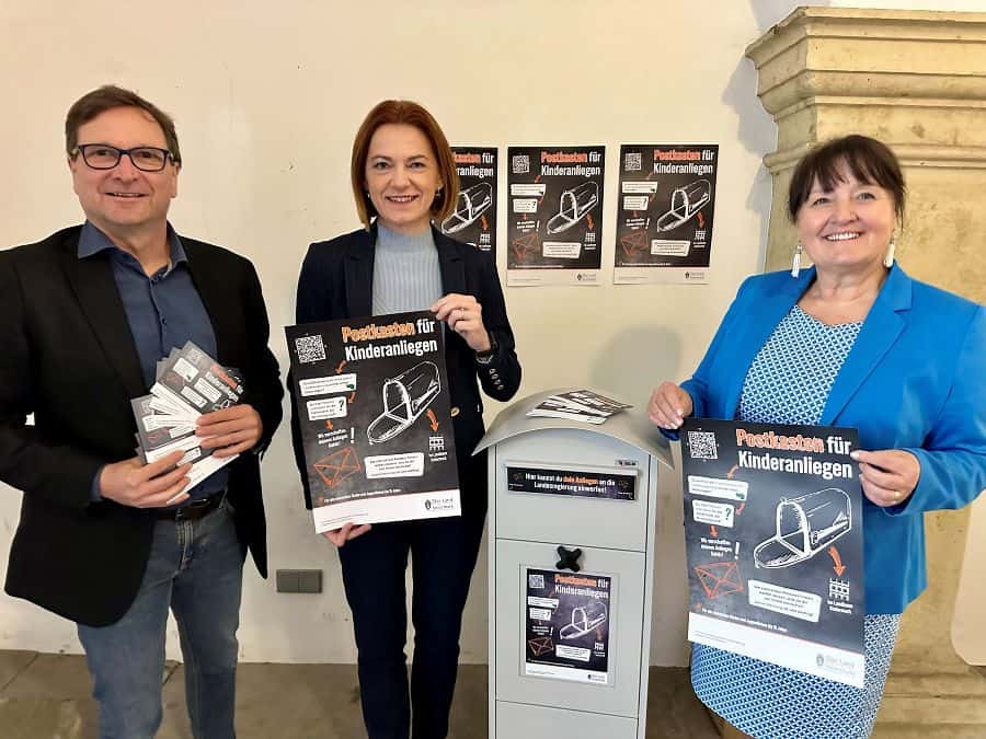 Bildbeschreibung: Zwei Frauen und ein Mann stehen neben einem Briefkasten und halten Infomaterial in ihren Händen.