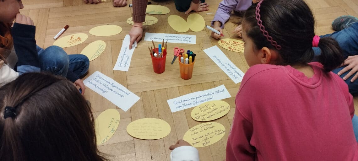 Kinder von hinten am Bodensitzend beim beantworten von Fragekärtchen zum Thema Schulessen
