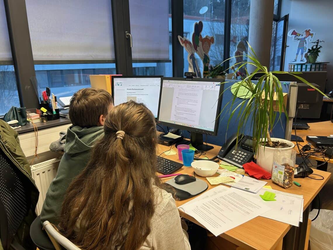 zwei Kinder von hinten, die an einem Schreibtisch sitzen und etwas am Computer arbeiten