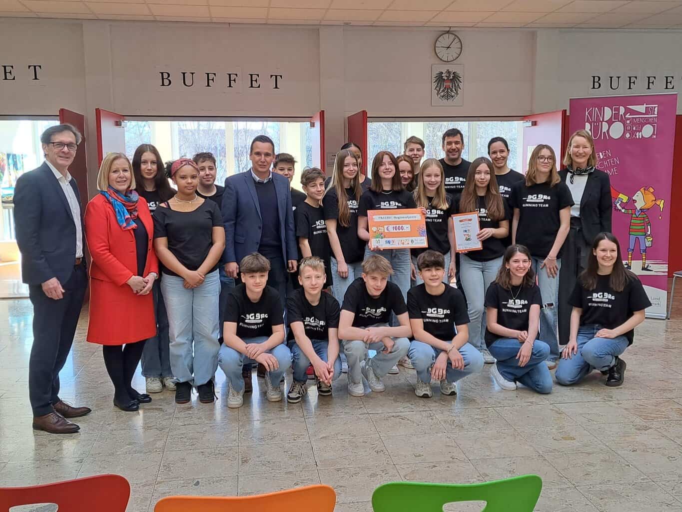 eine Gruppe Schüler*innen in schwarzen T-Shirts und ein paar Erwachsene an der Seite, alle schauen in die Kamera in der Mitte hält ein Mädchen einen Preisscheck hoch