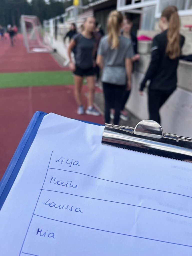 ein Klemmbrett, auf dem Namen stehen, im Hintergrund eine Gruppe Mädchen auf einem Sportplatz