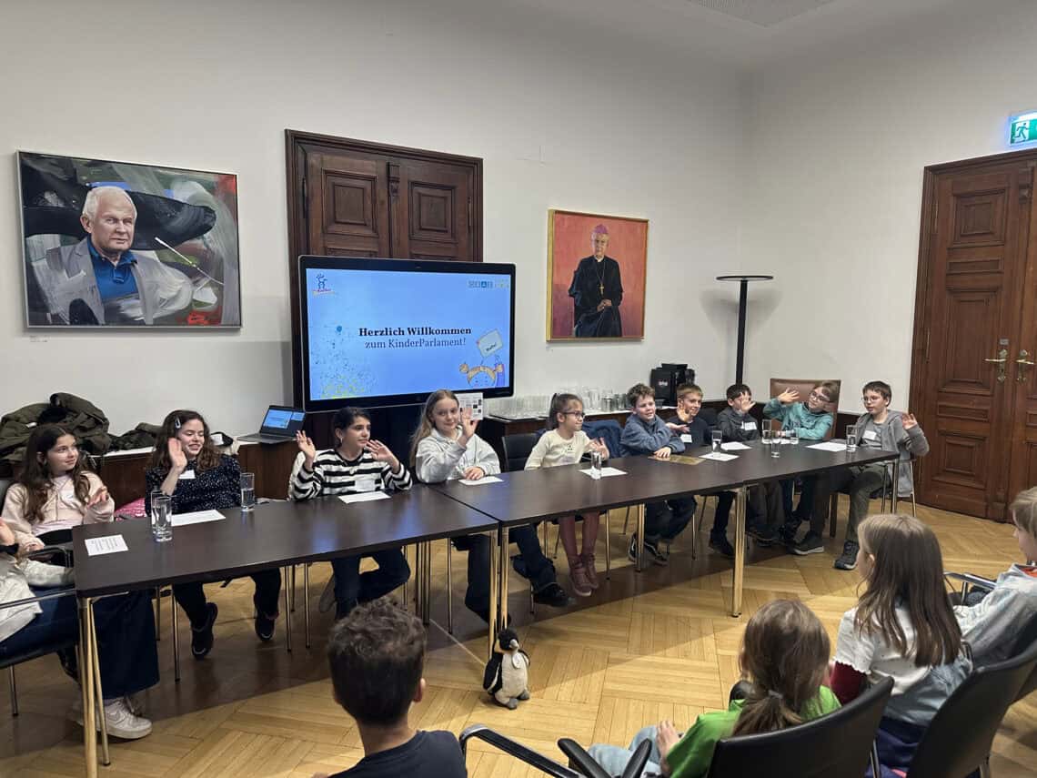 Eine Gruppe Kinder, die an einem langen Tisch sitzt und miteinander redet, im Hintergrund ist ein Bildschirm "Willkommen im KinderParlament"!