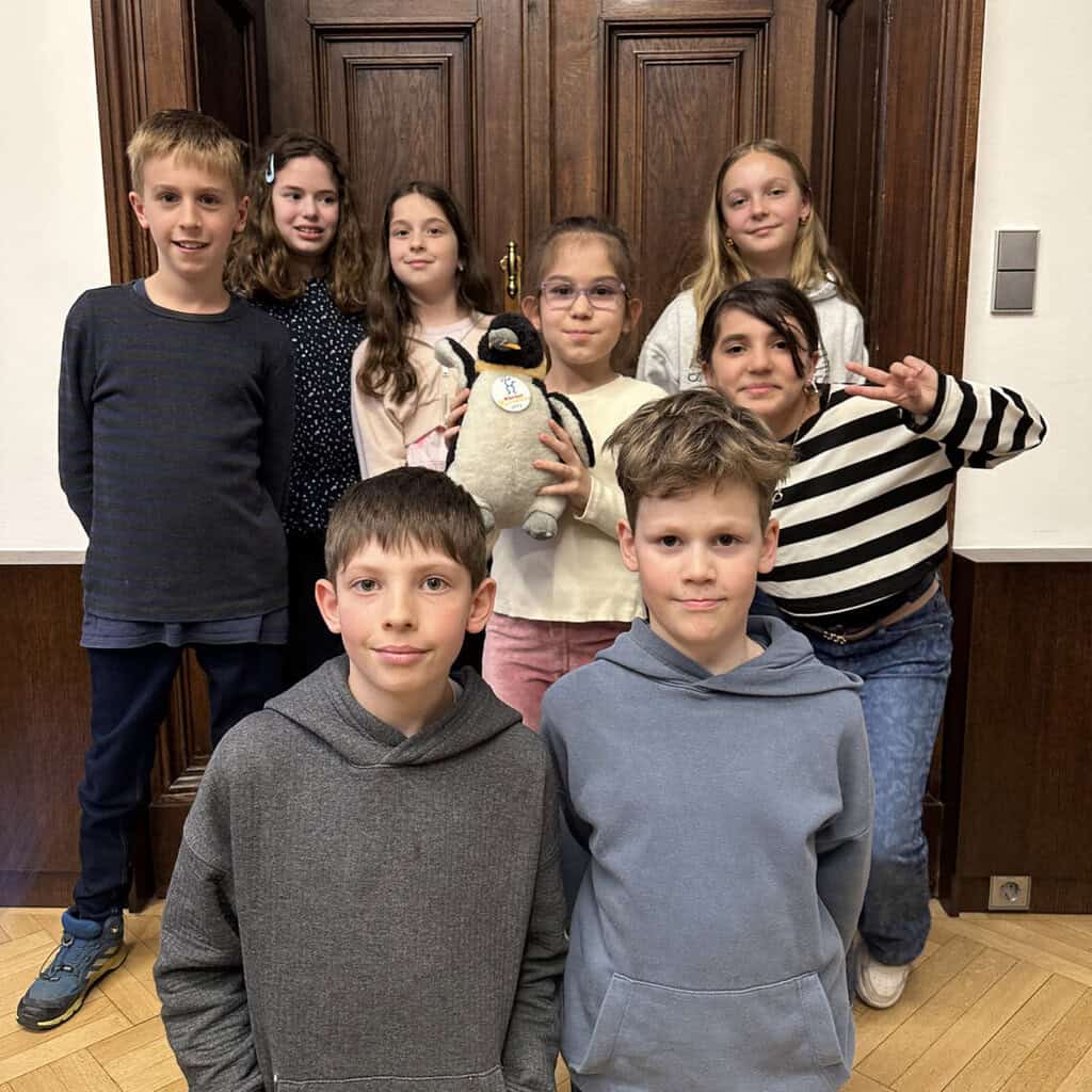eine Gruppe Kinder, die in einem Innenraum vor einer braunen Holztür steht. Ein Mädchen in der Mitte hält einen Plüschpinguin in die Kamera.